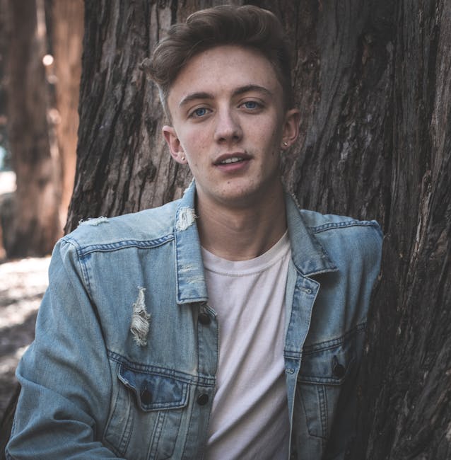 Casual portrait of a young man in a denim jacket leaning against a tree.
